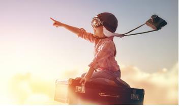 Child with helmet, goggles and camera flying in the clouds sitting on a suitcase while pointing and imaging success in life.
